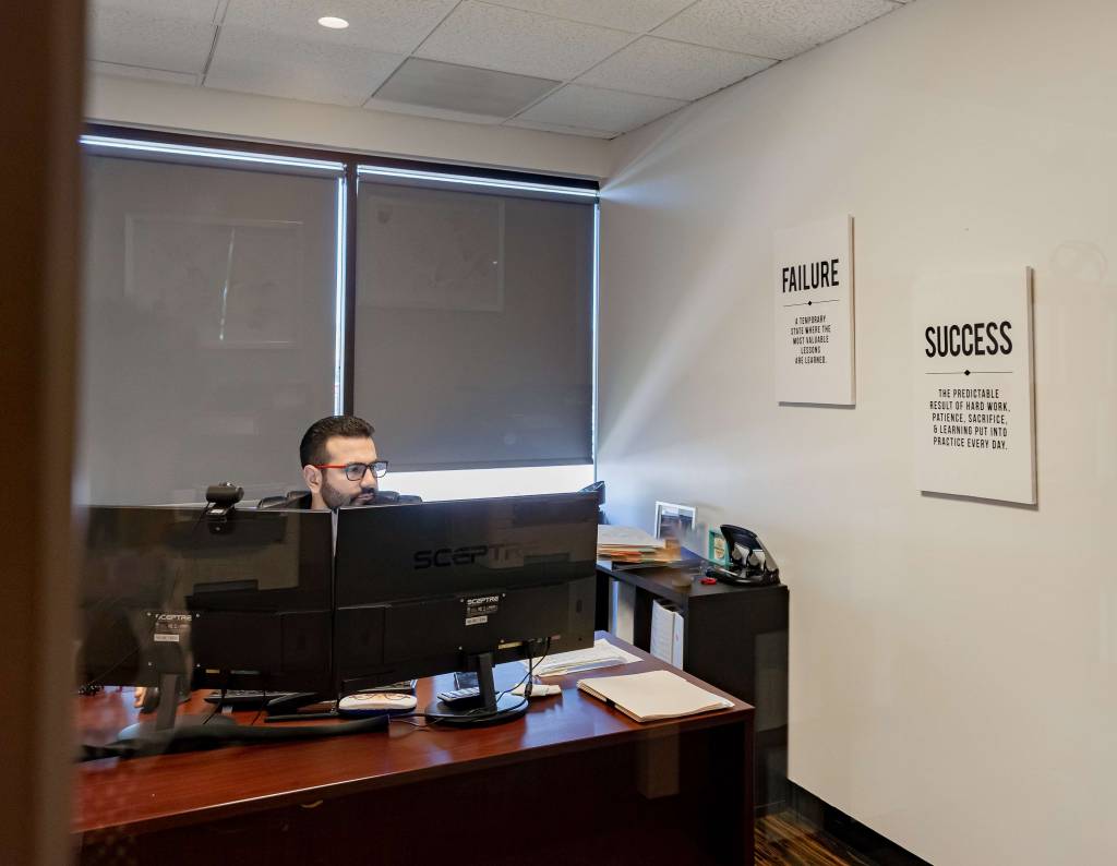 A man with glasses working at a desk with dual computer monitors and motivational office wall art.